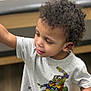 child, toddler, curly_hair, gray_tshirt, cartoon_print, teenage_mutant_ninja_turtles, indoor, wooden_floor, smiling, face, arm, short_sleeve, cute, young, person, casual_clothing, expression, playful, looking_away, skin