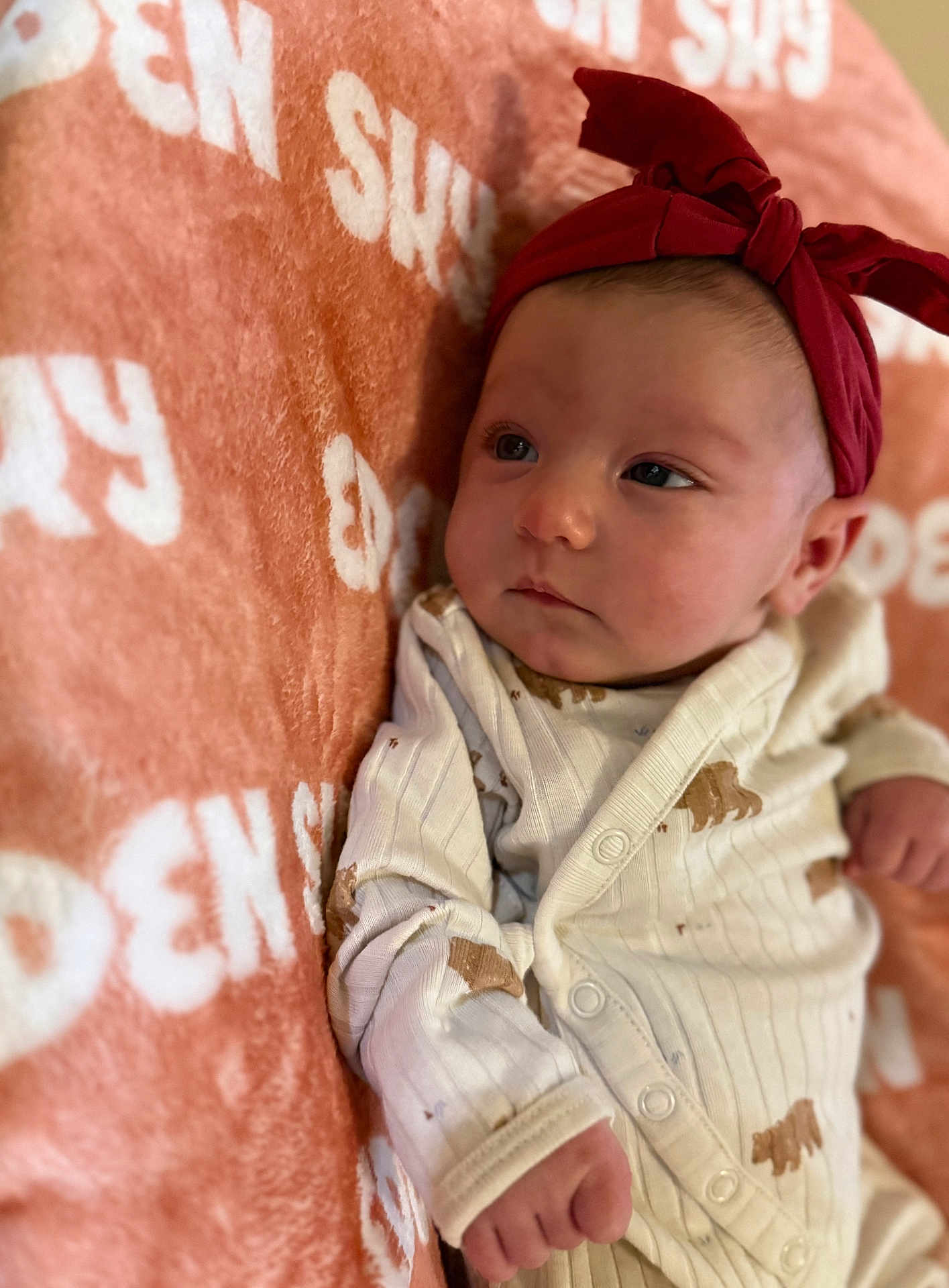 Eden is registered to the contest to win money with this photo: baby, infant, headband, red_bow, blanket, orange_blanket, white_text, onesie, bear_pattern, clothing, face, skin, hand, portrait, resting, indoor, soft_texture, cute, newborn, child