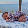 baby, sleeping, plaid_shirt, blanket, outdoor, beach, water, tree_trunk, peaceful, infant, soft_texture, blue_sky, nature, cute, child, portrait, relaxation, calm, sock, toddler