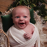 Hunter is registered to the contest to win money with this photo: baby, smiling, blanket, basket, holly_leaves, curly_fur, wood_floor, green_pillow, cute, infant, wrapped, portrait, happy, cozy, soft_texture, indoors, child, face, newborn, warm