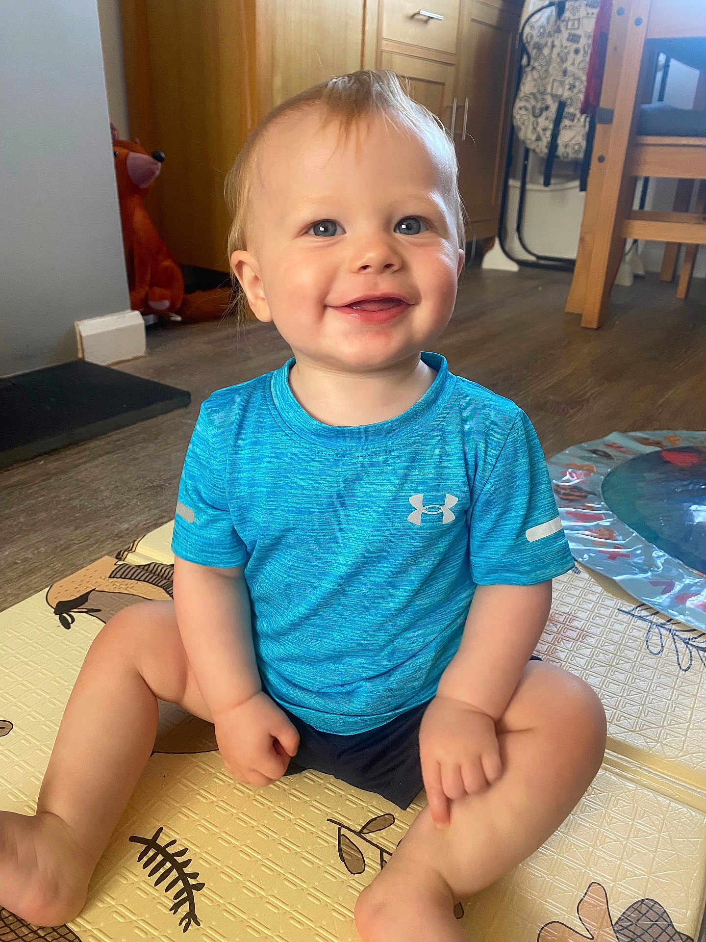 Hunter is registered to the contest to win money with this photo: toddler, child, smiling, blue_shirt, indoor, play_mat, cross_legged, floor, wooden_furniture, plush_toy, happy, cute, person, baby, young_child, short_hair, blue_eyes, casual_clothing, home, flooring
