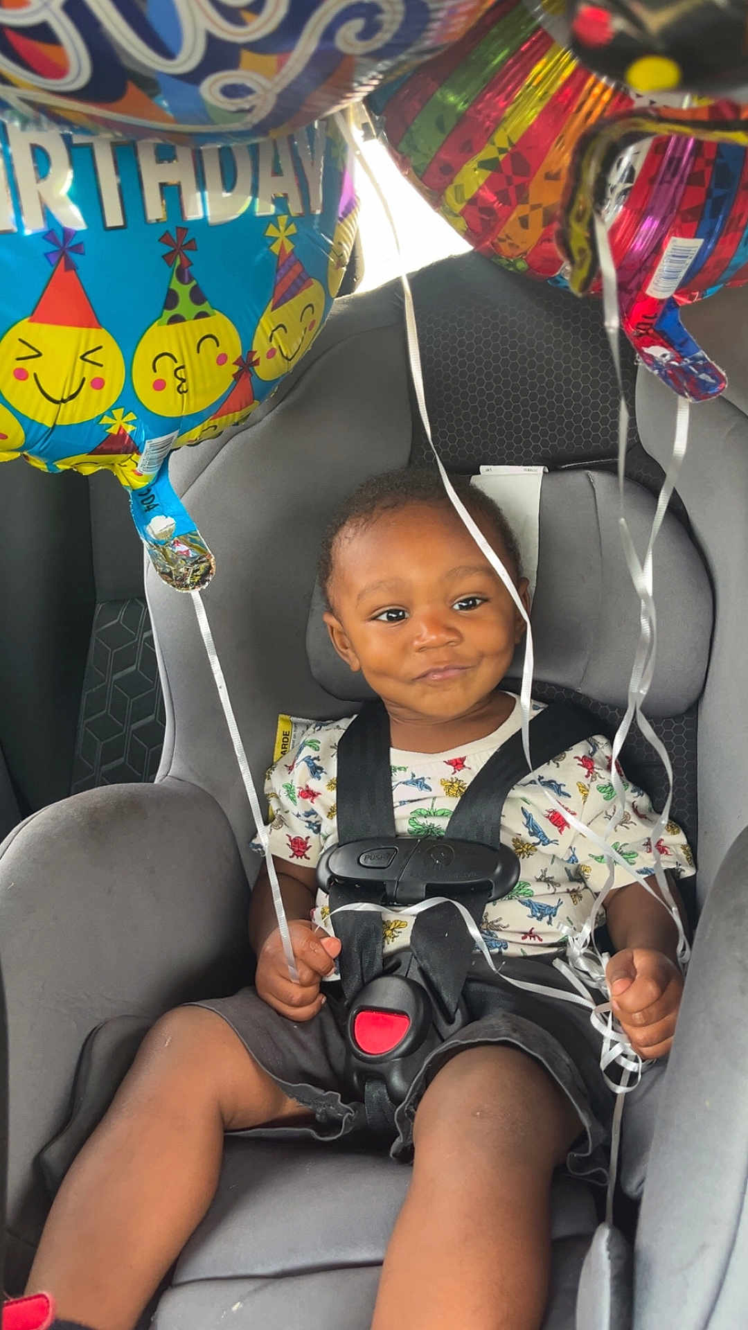 Kingsley is registered to the contest to win money with this photo: toddler, child, car_seat, birthday_balloons, smiling, harness, dinosaur_shirt, seatbelt, car_interior, celebration, party, colorful, happy, infant, young_child, sitting, vehicle, strapped_in, cute, ribbons