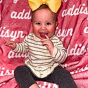 Addisyn joined the competition — help win amazing prizes! baby, child, smiling, yellow_bow, striped_sweater, dark_leggings, pink_blanket, text_background, sitting, happy, cute, indoors, portrait, face, finger_in_mouth, infant, young_child, bright_colors, cozy, playful
