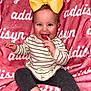 baby, child, smiling, yellow_bow, striped_sweater, dark_leggings, pink_blanket, text_background, sitting, happy, cute, indoors, portrait, face, finger_in_mouth, infant, young_child, bright_colors, cozy, playful