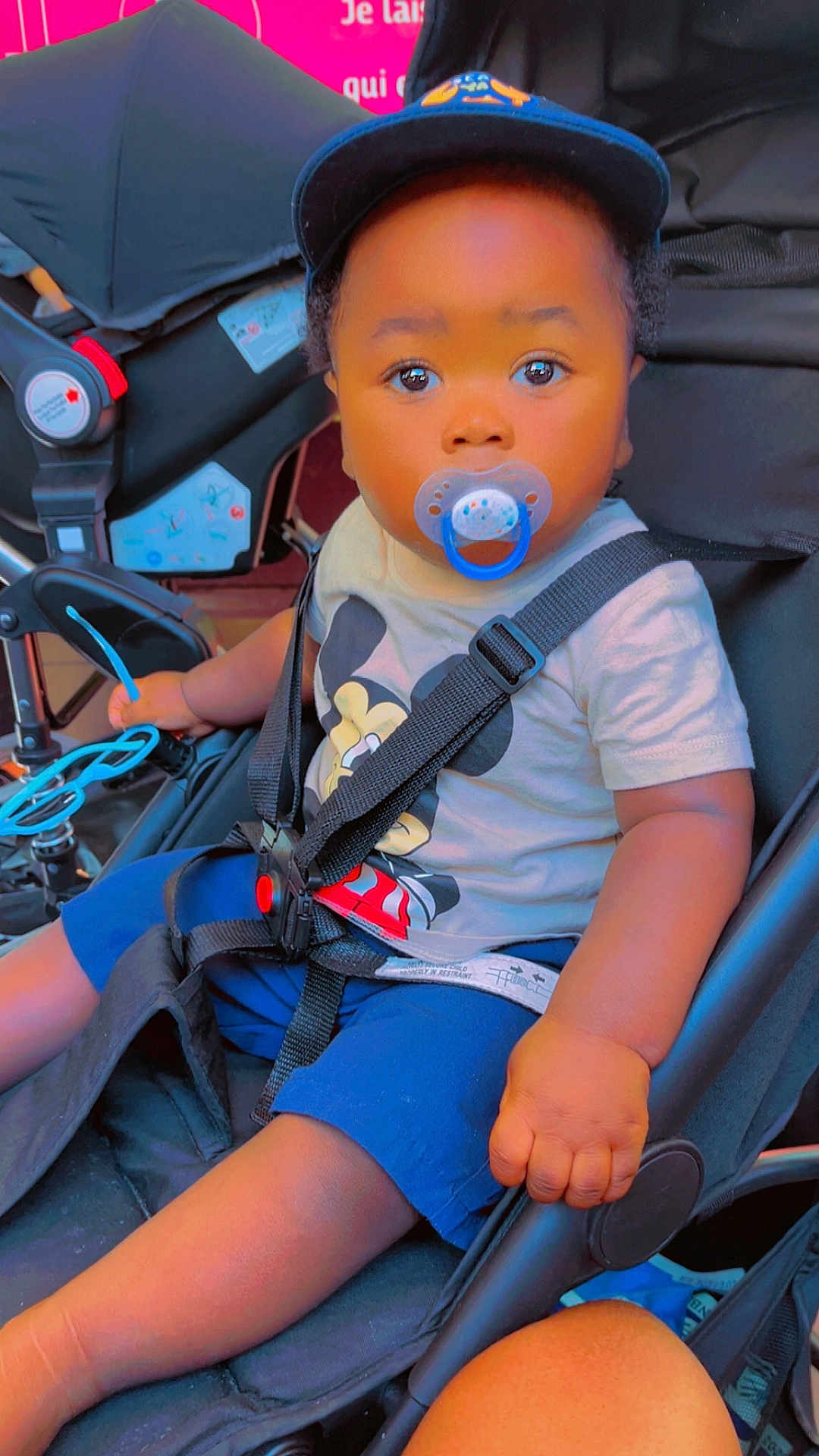 Lucas-Joao Zayn participe au concours pour gagner de l'argent avec cette photo : toddler, child, pacifier, stroller, cap, mickey_mouse, shirt, shorts, seatbelt, hand, face, sitting, blue, toy, baby, infant, young_child, cute, outdoor, baby_clothing