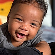 Benjamin is registered to the contest to win money with this photo: baby, smile, face, child, curly_hair, black_mitten, grey_clothing, blanket, indoor, portrait, cute, happy, infant, closeup, soft_texture, warm_lighting, background_blur, person, toddler, expression