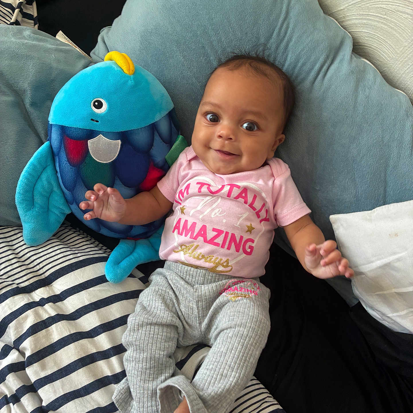 Amazing is registered to the contest to win money with this photo: baby, child, plush_toy, fish_toy, pillow, bed, striped_bed_sheet, pink_shirt, gray_pants, smiling, happy, indoors, person, infant, cute, cozy, soft_texture, portrait, lying_down, casual_clothing