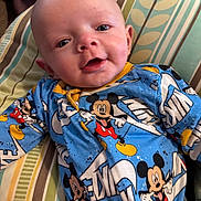 Oliver is registered to the contest to win money with this photo: baby, infant, mickey_mouse, onesie, clothing, pillow, striped_pillow, blue, yellow, red, smile, face, head, person, cute, indoor, child, young, happy, lying_down