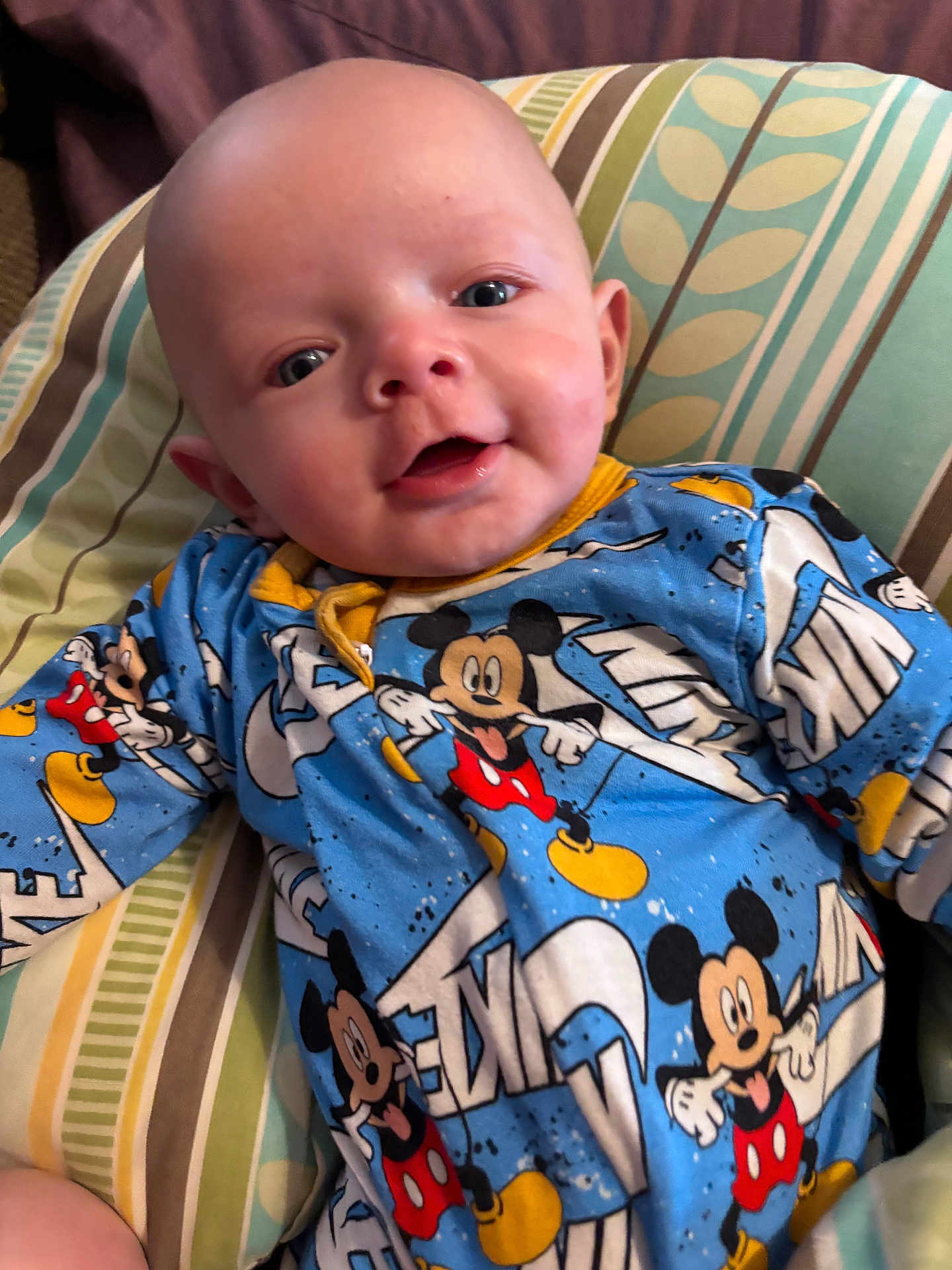 Oliver is registered to the contest to win money with this photo: baby, infant, mickey_mouse, onesie, clothing, pillow, striped_pillow, blue, yellow, red, smile, face, head, person, cute, indoor, child, young, happy, lying_down