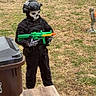 child, toy_gun, skeleton_mask, black_clothing, helmet, gloves, backyard, grass, trash_bin, wooden_furniture, imaginative_play, costume, outdoor, person, toy, mask, helmet_accessory, green_toy, orange_tip, play