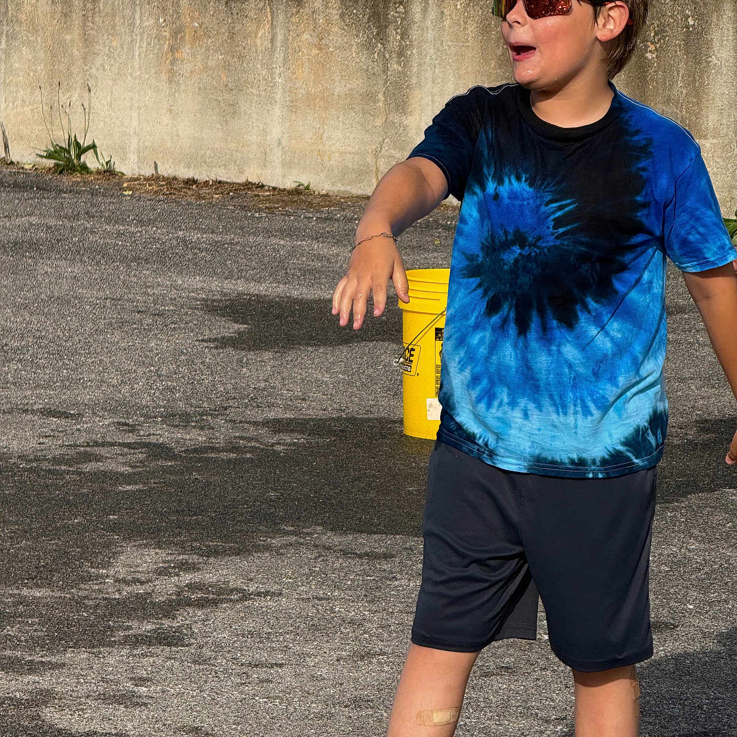 Malaki is registered to the contest to win money with this photo: active, bandage, blue_shirt, boy, casual_clothing, chain_link_fence, child, concrete_wall, fun, outdoor, pavement, playful, shadow, shorts, summer, sunglasses, sunlight, tie_dye, yellow_bucket, young