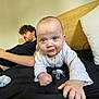 baby, man, bed, black_bedspread, pillow, pacifier, crawling, curly_hair, blue_eyes, white_clothing, headboard, indoor, relaxed, person, child, portrait, family, casual, furniture, cozy