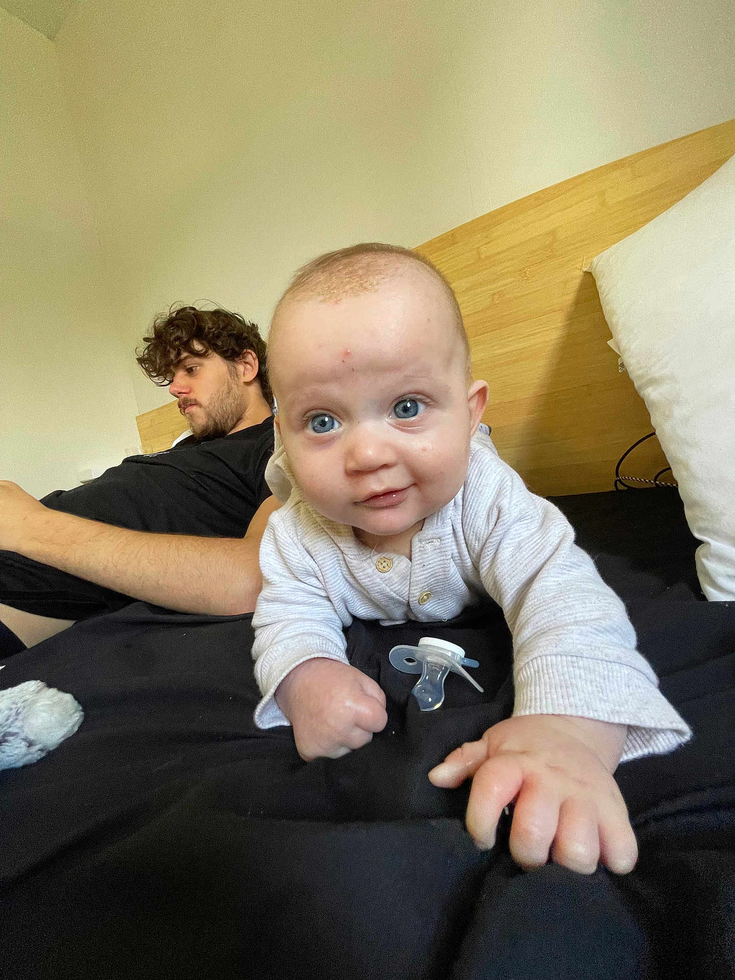 Lyam participe au concours pour gagner de l'argent avec cette photo : baby, man, bed, black_bedspread, pillow, pacifier, crawling, curly_hair, blue_eyes, white_clothing, headboard, indoor, relaxed, person, child, portrait, family, casual, furniture, cozy