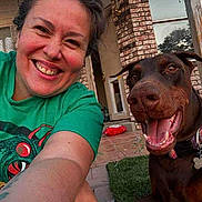 Gunner is registered to the contest to win money with this photo: woman, dog, doberman, smiling, selfie, outdoor, porch, brick_wall, grass, green_tshirt, happy, pet, collar, tongue_out, face, person, canine, sunlight, casual, joy