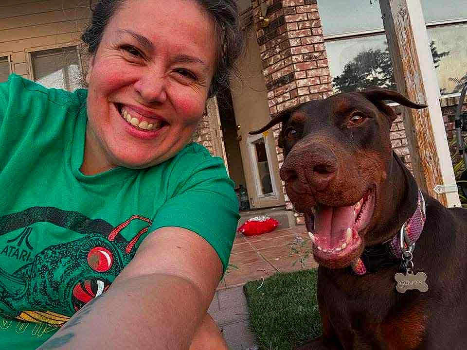 Gunner is registered to the contest to win money with this photo: woman, dog, doberman, smiling, selfie, outdoor, porch, brick_wall, grass, green_tshirt, happy, pet, collar, tongue_out, face, person, canine, sunlight, casual, joy
