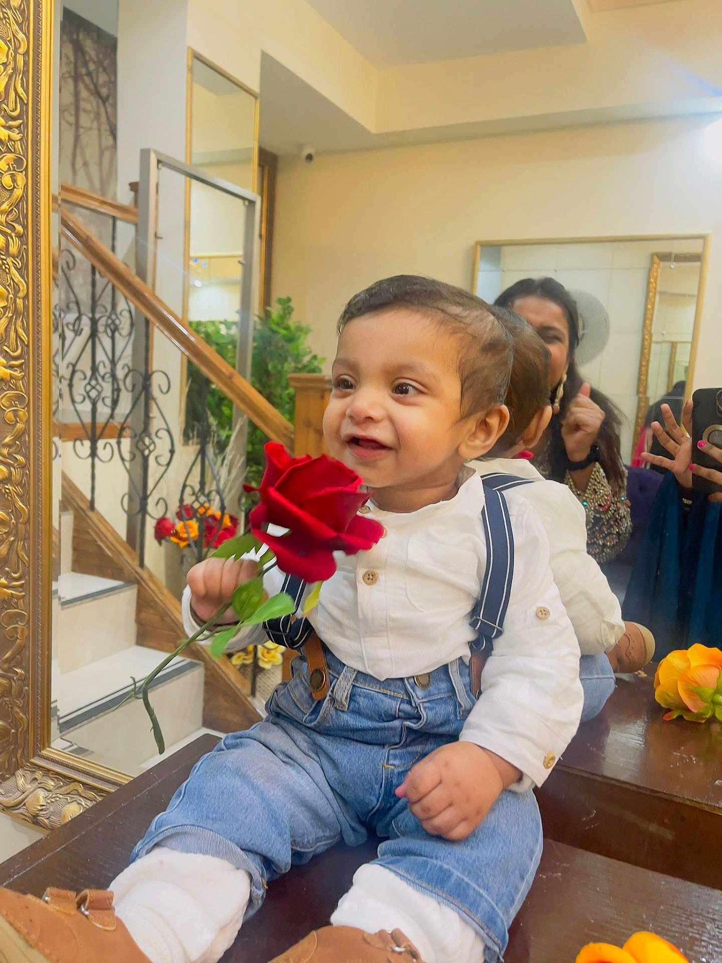 Gursher Singh is registered to the contest to win money with this photo: toddler, child, rose, flower, mirror, reflection, stairs, suspenders, jeans, white_shirt, smile, indoor, wooden_surface, decor, adult, thumbs_up, happy, casual_clothing, person, portrait