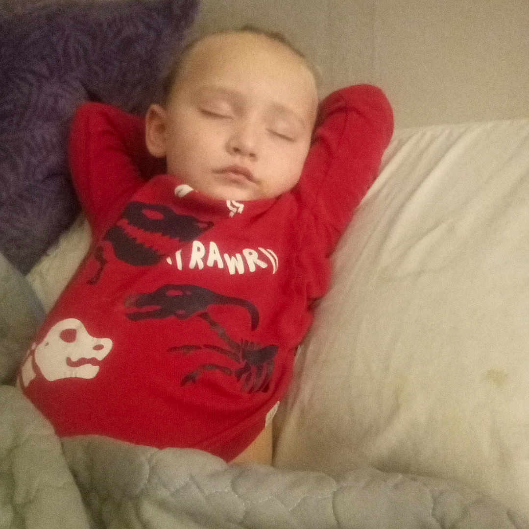 Myi-Khel is registered to the contest to win money with this photo: child, sleeping, bed, pillow, blanket, quilt, red_shirt, dinosaur, indoors, cozy, resting, peaceful, young, toddler, portrait, person, clothing, head, face, relaxation