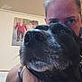 dog, person, selfie, close_up, black_dog, indoor, portrait, face, animal, pet, wall, photo_frame, human, eyes, snout, fur, casual_clothing, bracelet, background, home