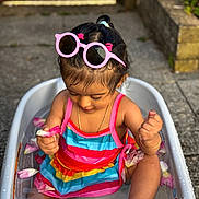 Heather is registered to the contest to win money with this photo: toddler, child, water, bathtub, flower_petals, pink_sunglasses, rainbow_striped_clothing, outdoor, summer, playful, happy, sitting, skin, hair, foot, hand, gold_bracelet, gold_chain, concrete, sunlight
