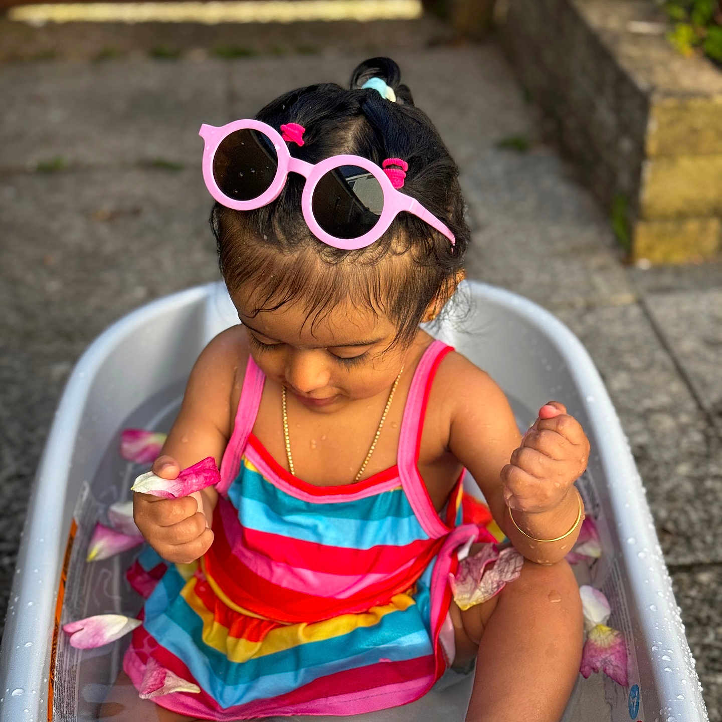 Heather is registered to the contest to win money with this photo: bathtub, child, concrete, flower_petals, foot, gold_bracelet, gold_chain, hair, hand, happy, outdoor, pink_sunglasses, playful, rainbow_striped_clothing, sitting, skin, summer, sunlight, toddler, water