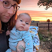 Fenn joined the competition — help win amazing prizes! baby, woman, glasses, tattoo, sunset, outdoor, blanket, floral_pajamas, field, fence, tree, sky, portrait, person, holding, nature, grass, face, expression, warm_light