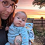 baby, woman, glasses, tattoo, sunset, outdoor, blanket, floral_pajamas, field, fence, tree, sky, portrait, person, holding, nature, grass, face, expression, warm_light