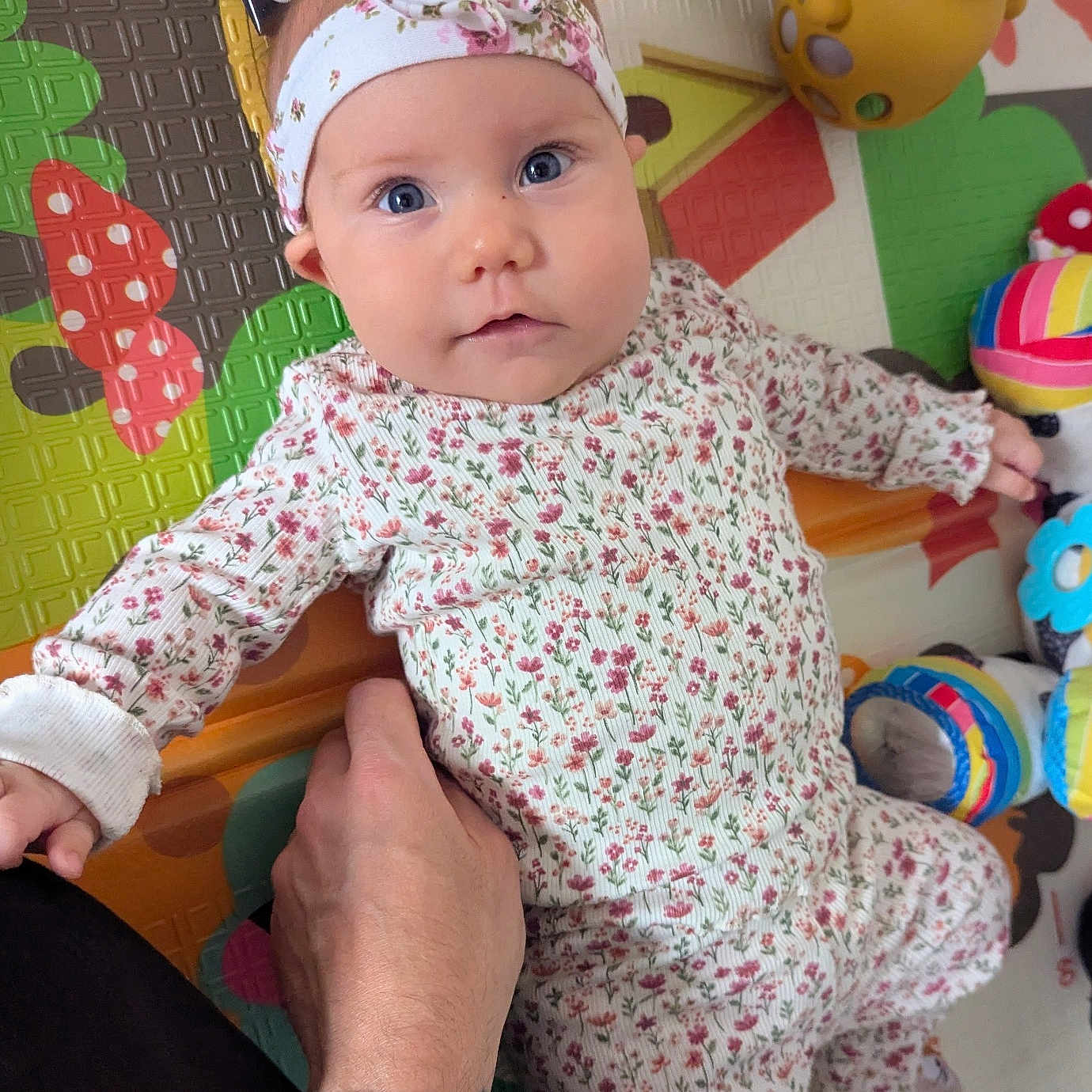 Fenn joined the competition — help win amazing prizes! baby, blue_eyes, child, clothing, colorful, cute, face, floral_pattern, hand, headband, indoor, infant, looking_at_camera, lying_down, person, play_mat, skin, sleeves, supportive_hand, toy