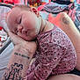 baby, sleeping, floral_onesie, tattoo, arm, hand, person, indoors, blurred_background, soft_lighting, skin, face, peaceful, closeup, child, cozy, resting, furniture, bed, pink_tone