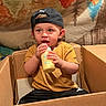 Pierston is registered to the contest to win money with this photo: baby_bottle, black_cap, blanket, cap, cardboard_box, casual_clothing, child, colorful_background, cute, drinking, expression, face, indoor, mustard_shirt, person, portrait, relaxed, sitting, toddler, young_child