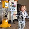 boots, casual_clothing, child, cute, floor, gumball_machine, happy, indoor, patterned_clothing, person, playful, real_photo, smiling, sweatpants, toddler, toy, vending_machine, walking, yellow, young_child