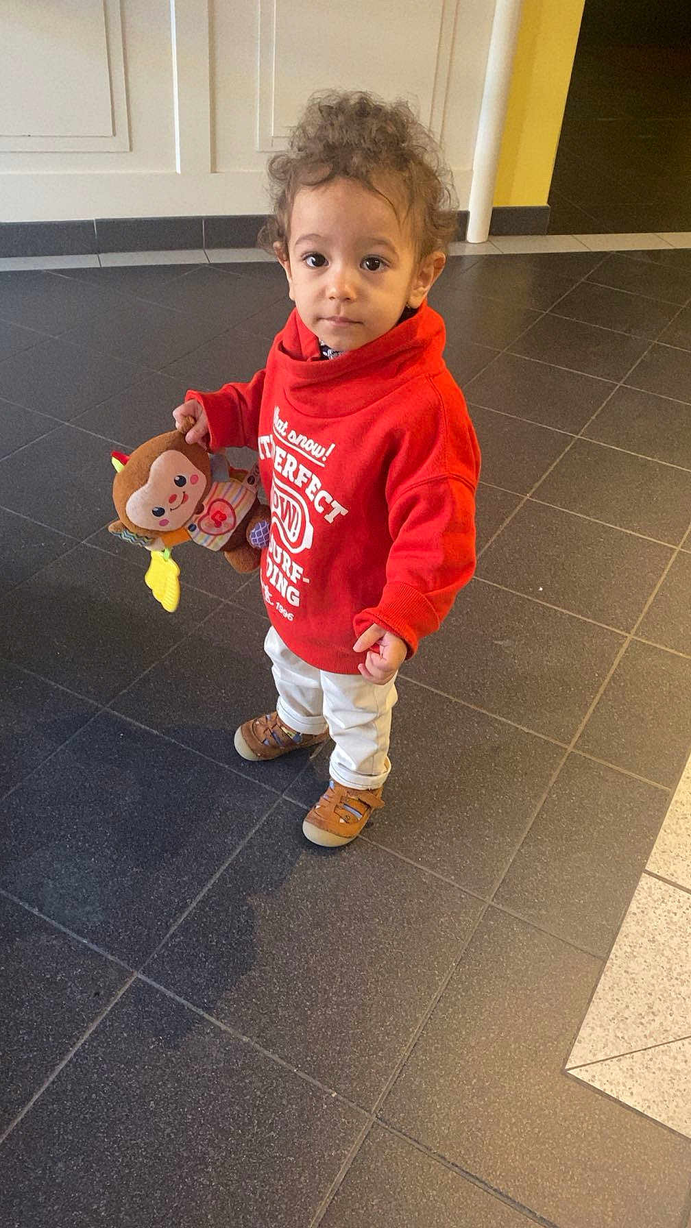 Ilyan a rejoint le concours — aidez-le/la à gagner de superbes lots ! toddler, child, indoor, toy, stuffed_animal, curly_hair, red_sweatshirt, white_pants, brown_shoes, floor_tiles, person, young_child, looking_at_camera, cute, casual_clothing, playful, expression, standing, holding_toy, curious