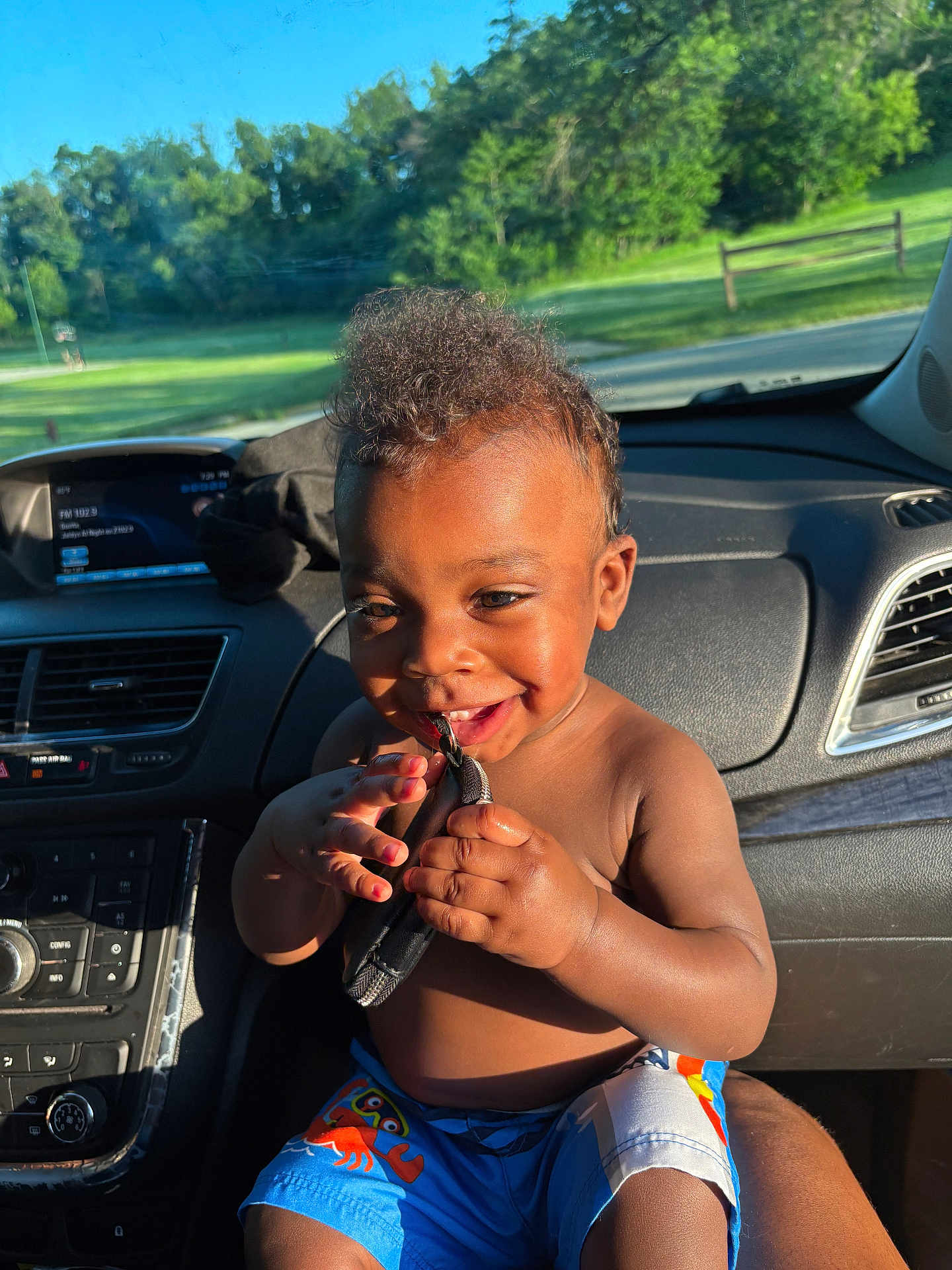 Kaysyn is registered to the contest to win money with this photo: toddler, child, car_interior, sunlight, smiling, shorts, blue_shorts, crab_pattern, hands, face, happy, playful, seat, dashboard, window, trees, greenery, daylight, skin, person