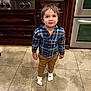 toddler, child, kitchen, floor, tile, plaid_shirt, pants, socks, wooden_cabinets, stainless_steel_appliance, oven, stove, curious, standing, indoor, young_child, brown_hair, smiling, casual_clothing, home