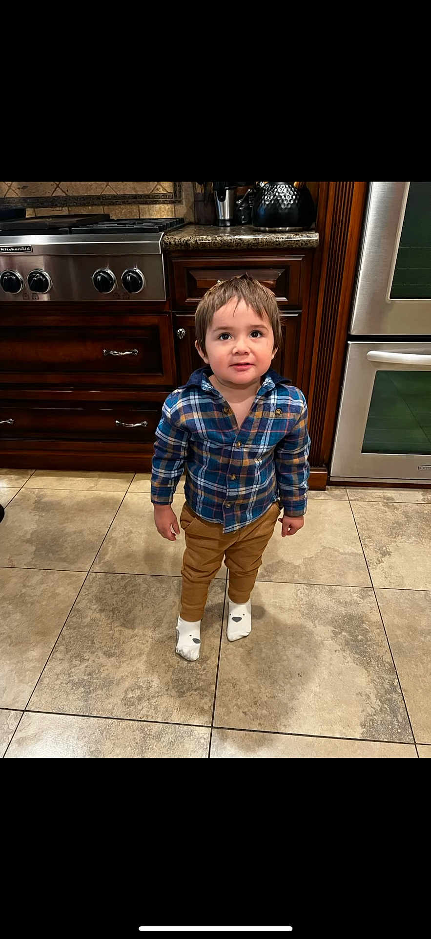 Calvin joined the competition — help win amazing prizes! toddler, child, kitchen, floor, tile, plaid_shirt, pants, socks, wooden_cabinets, stainless_steel_appliance, oven, stove, curious, standing, indoor, young_child, brown_hair, smiling, casual_clothing, home