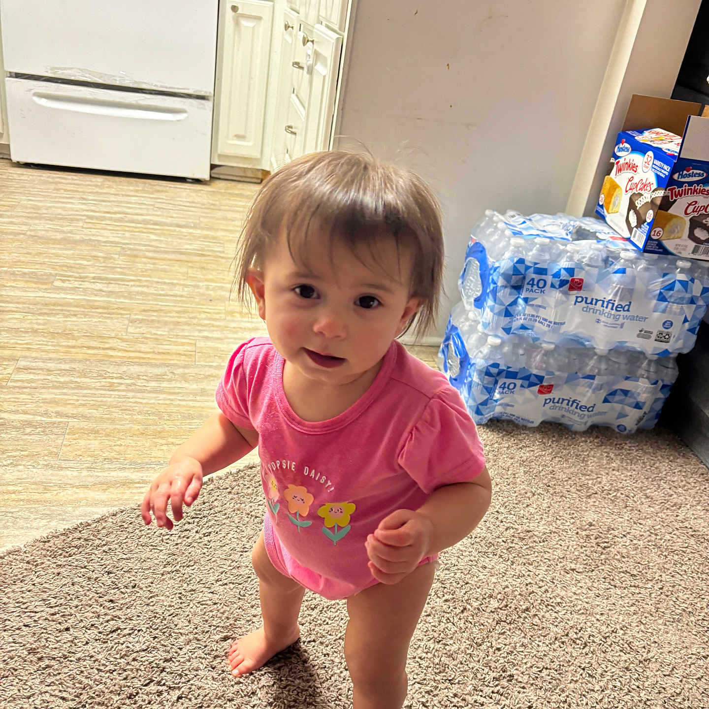 Alisia Buccheri joined the competition — help win amazing prizes! barefoot, cabinet, carpet, child, curious, expression, face, floor, hair, home, indoors, kitchen, person, pink_onesie, snack_boxes, standing, stove, toddler, water_bottles, young_child