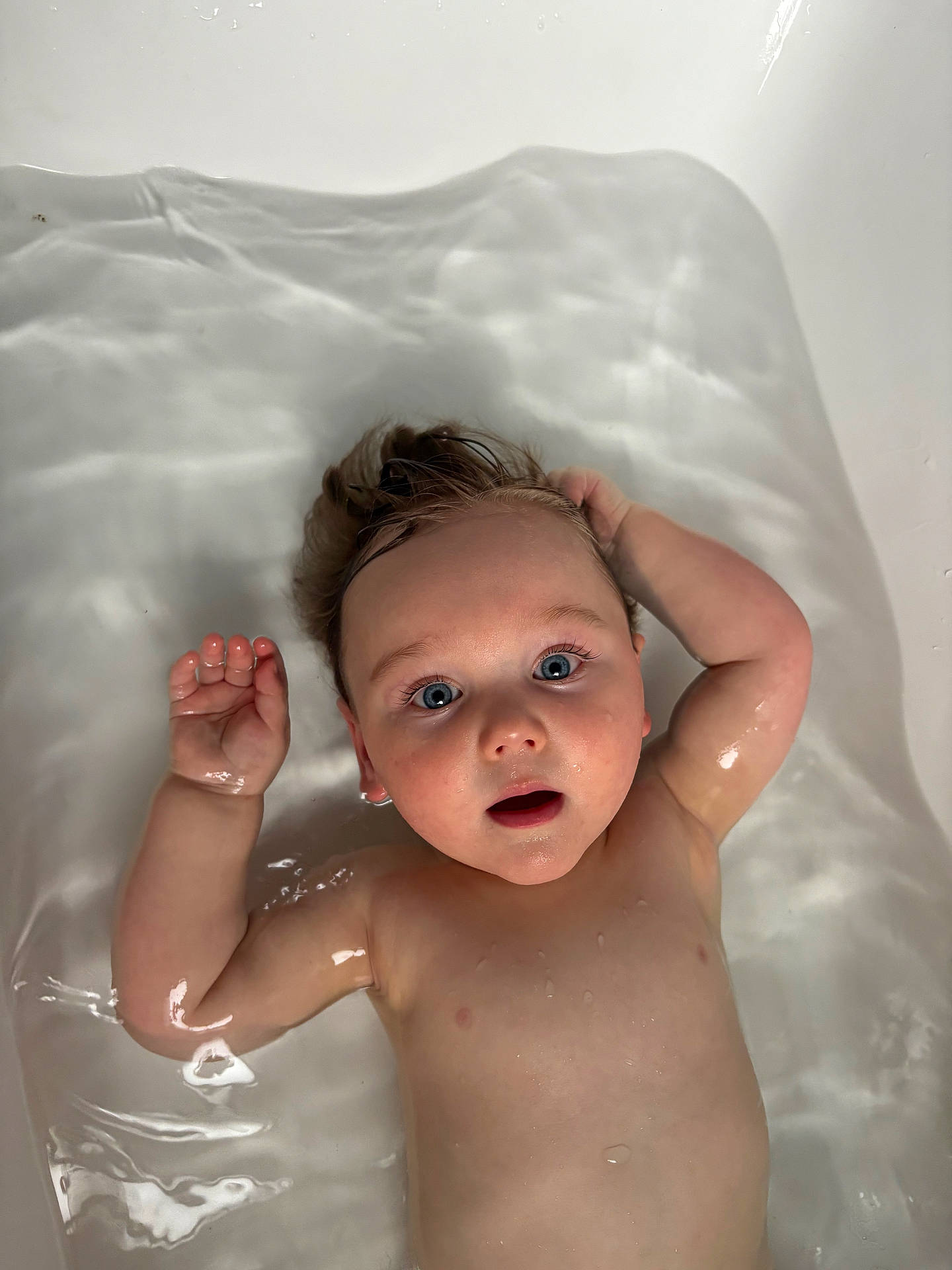 arm, baby, bath_time, bathtub, blue_eyes, child, curly_hair, expression, face, hand, head, indoors, infant, nude, person, reflection, skin, water, wet, young