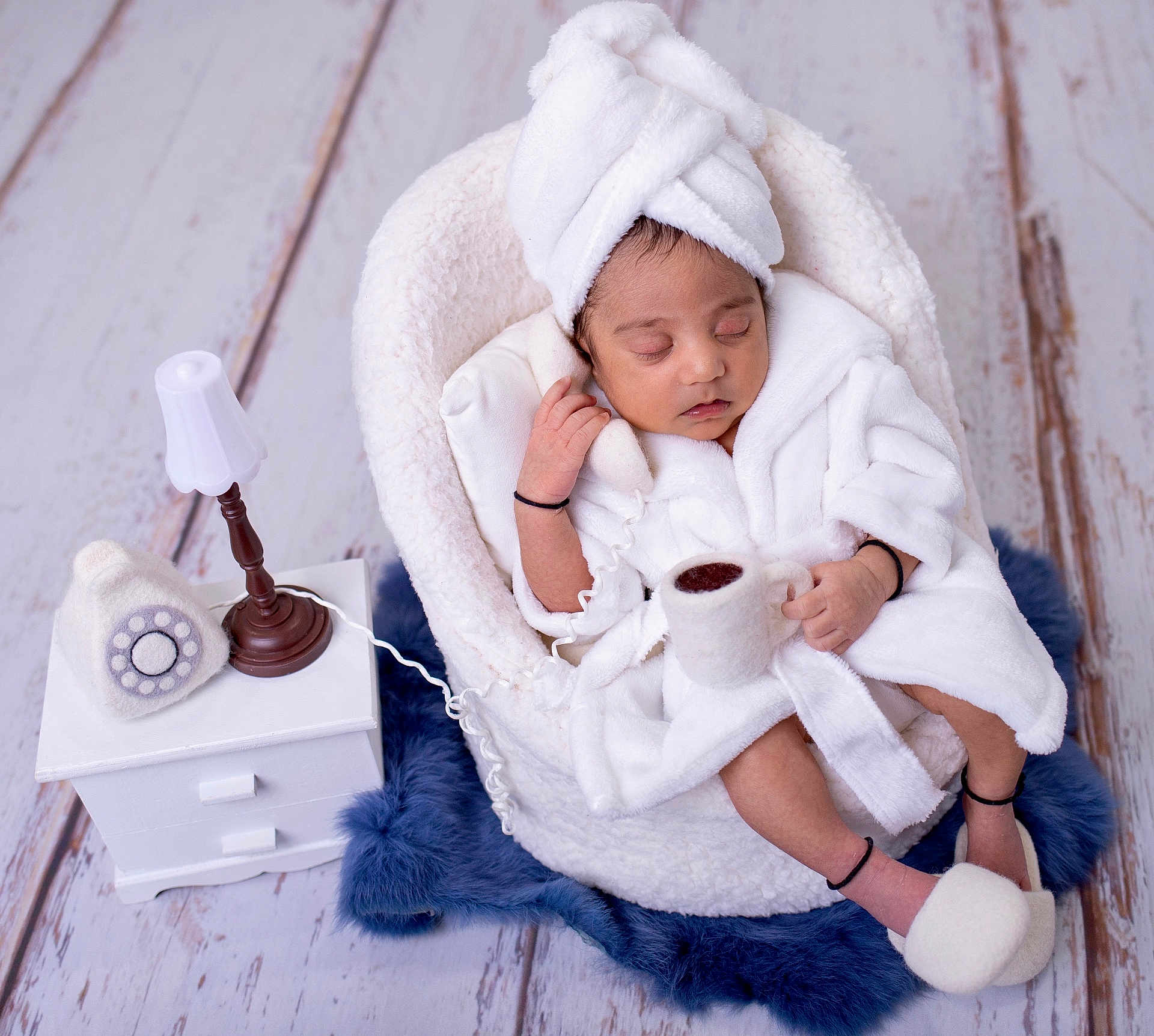 Aarav is registered to the contest to win money with this photo: newborn, baby, sleeping, white_robe, towel_turban, basket, cushion, blue_rug, floor, miniature_lamp, rotary_phone, small_table, indoor, cozy, cute, peaceful, infant, soft_texture, relaxing, portrait