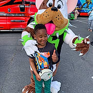 Jayden a rejoint le concours — aidez-le/la à gagner de superbes lots ! asphalt, child, costume, crossbody_bag, daylight, dog_costume, event, fun, green_hat, mascot, outdoor, parking_lot, person, red_truck, smile, sneakers, spiked_collar, t_shirt, thumbs_up, vehicle