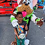 child, mascot, dog_costume, thumbs_up, parking_lot, red_truck, green_hat, spiked_collar, t_shirt, sneakers, crossbody_bag, person, outdoor, smile, costume, fun, event, vehicle, asphalt, daylight