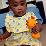 baby, child, toy, yellow_dress, purple_flowers, indoor, floor, curly_hair, dark_skin, sitting, cute, playful, hand, face, eyes, expression, home, room, furniture, diaper