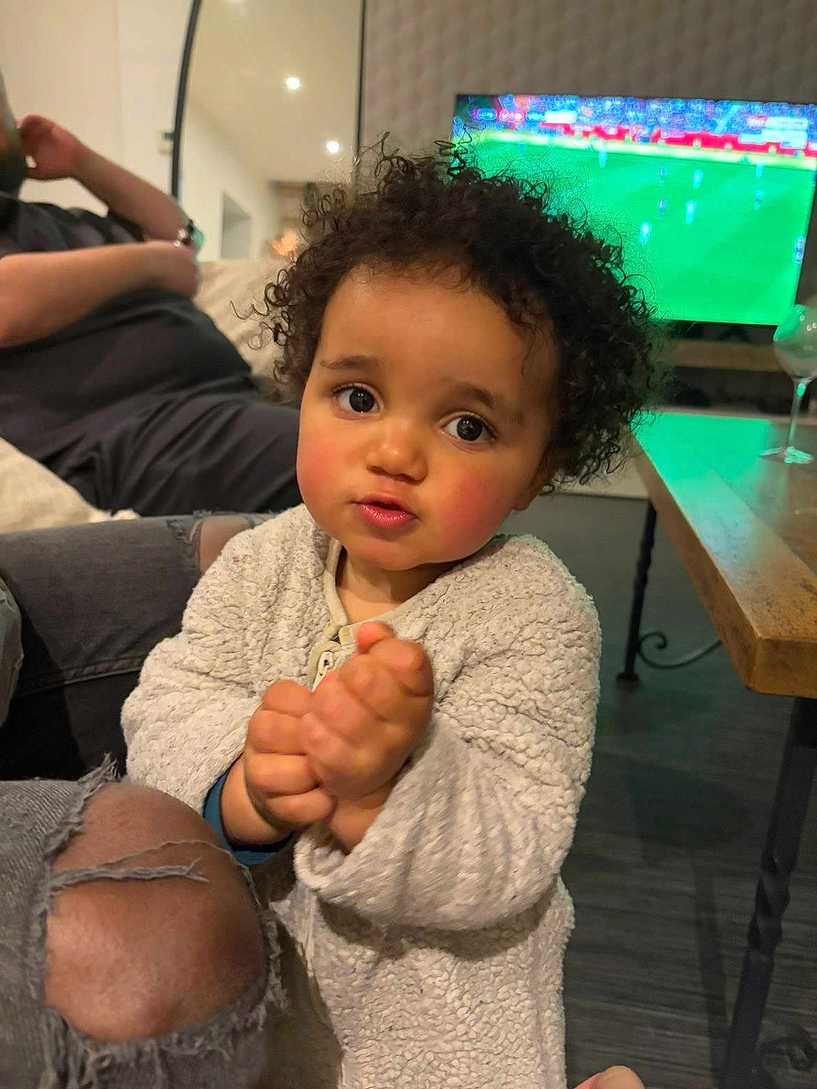 Eli participe au concours pour gagner de l'argent avec cette photo : child, toddler, curly_hair, big_eyes, clasped_hands, living_room, television, soccer, sofa, couch, ripped_jeans, wooden_table, wine_glass, person, cozy_clothing, flooring, portrait, indoor, cute, family_moment