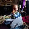 baby_seat, blanket, cabinet, child, curly_hair, dog, dolls, food, happy, home_interior, indoor, person, pink_cushion, plate, purple_curtain, shelf, smiling, snack, television, toddler