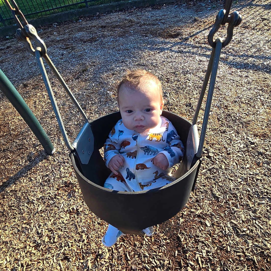 Lucius is registered to the contest to win money with this photo: animal_print, baby, bucket_seat, car, chain, expression, fence, house, infant, onesie, outdoors, park, playground, portrait, seat, shadow, shoe, sunlight, swing, wood_chips