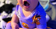 Bailey is registered to the contest to win money with this photo: baby, child, infant, portrait, sitting, wide_eyes, open_mouth, surprised, indoor, pajamas, cartoon_clothing, colorful, purple_lighting, blue_lighting, close_up, face, hands, toys, blanket, home