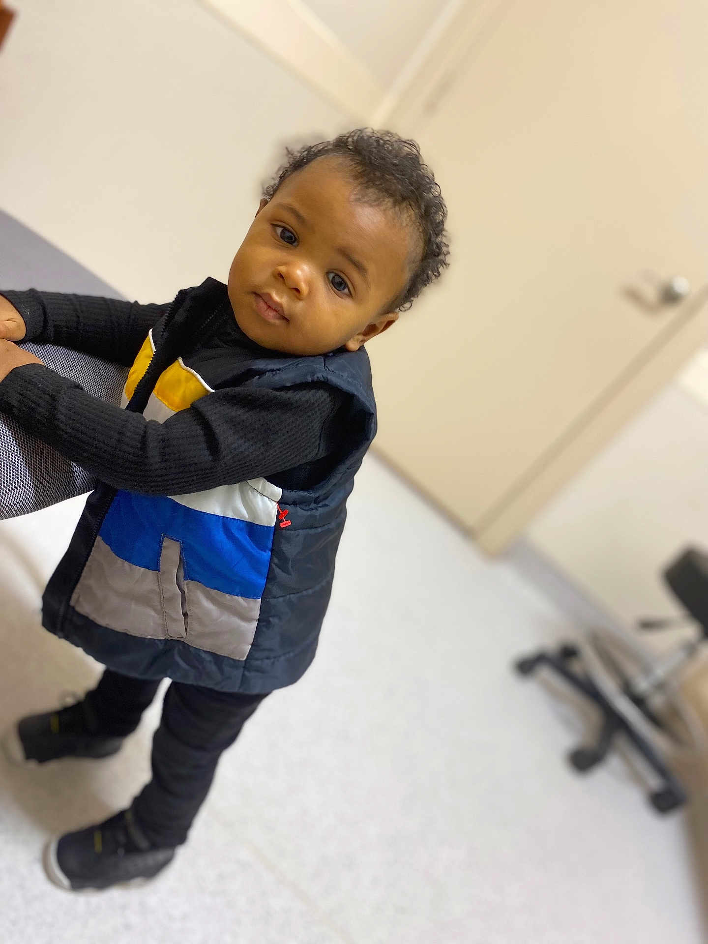 Messiah joined the competition — help win amazing prizes! toddler, child, indoor, curious, vest, black_clothing, standing, chair, floor, door, office_chair, portrait, person, young_child, footwear, looking, expression, casual, hair, skin