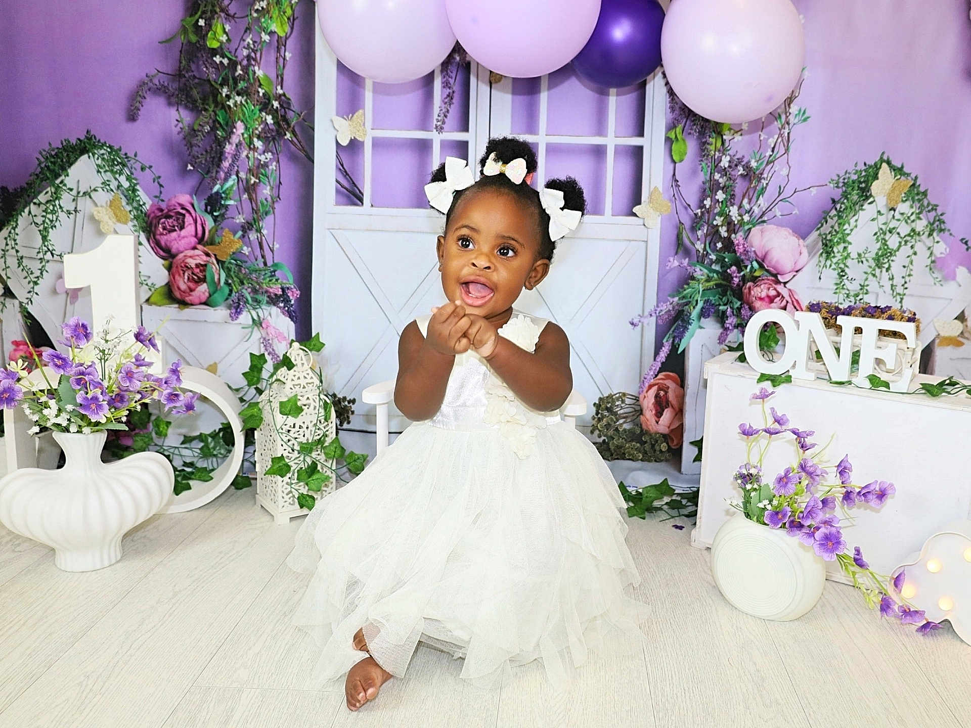 Mia is registered to the contest to win money with this photo: toddler, girl, white_dress, birthday, balloons, purple, flowers, decorations, smiling, happy, celebration, indoor, portrait, child, hair_bows, floor, props, party, cute, one_year_old