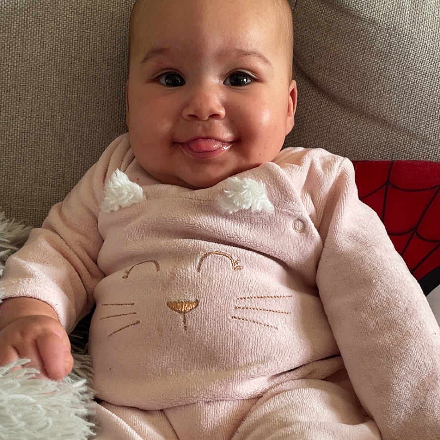 Halya participe au concours pour gagner de l'argent avec cette photo : baby, child, couch, cozy, cushion, cute, fabric, face, hand, home_interior, indoor, legs, onesie, photography, pillow, pink_outfit, portrait, sitting, smiling, tongue_out