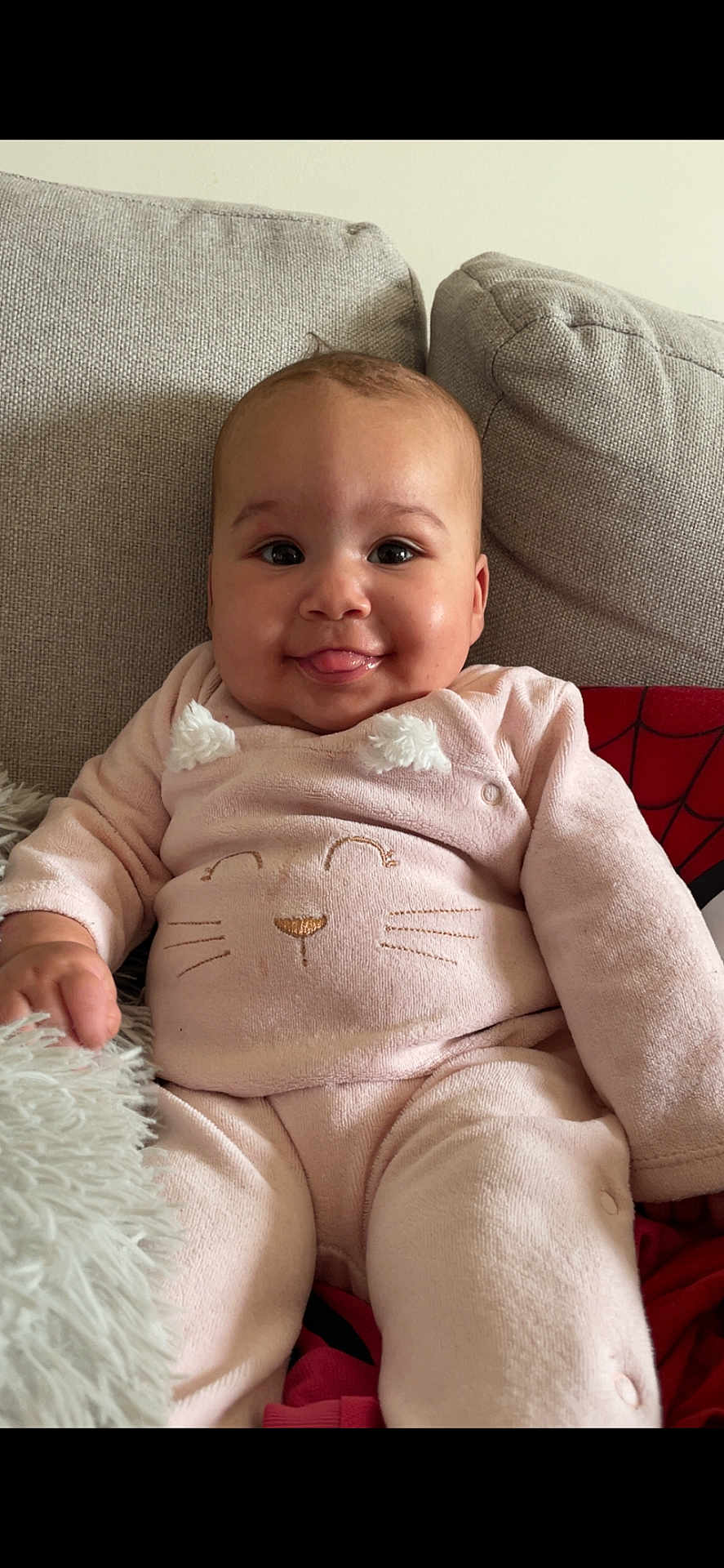 Halya participe au concours pour gagner de l'argent avec cette photo : baby, child, smiling, tongue_out, onesie, pink_outfit, couch, cushion, pillow, indoor, portrait, sitting, face, hand, legs, cozy, fabric, home_interior, cute, photography