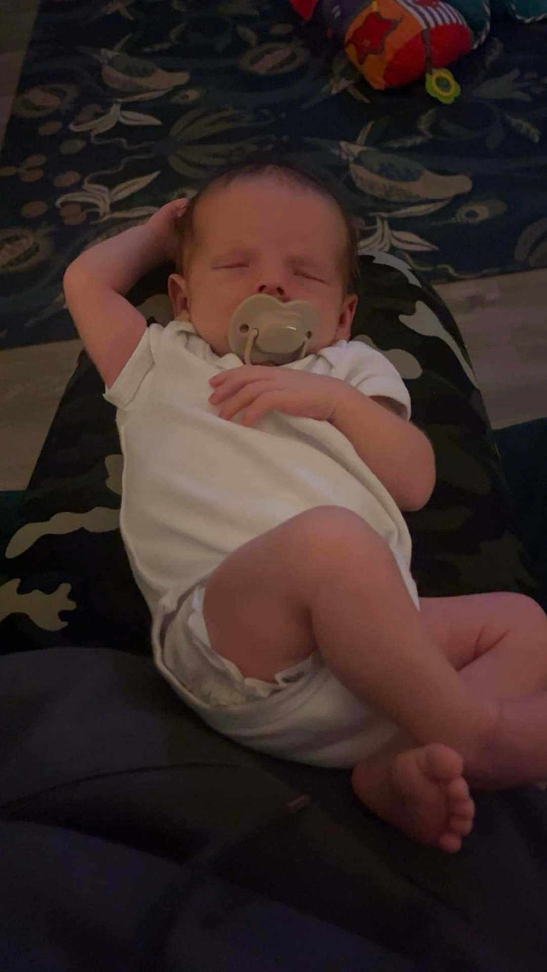 Jenson is registered to the contest to win money with this photo: baby, sleeping, pacifier, white_onesie, infant, resting, couch, cushion, hand, foot, person, indoor, comfort, relaxation, child, human, portrait, peaceful, nap, soft_lighting