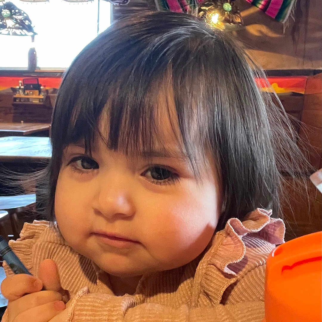 Luz joined the competition — help win amazing prizes! child, curly_hair, cute, decor, expression, face, hands, indoor, light, pen, person, pink_clothing, portrait, restaurant, sitting, table, toddler, window, wood, young_child